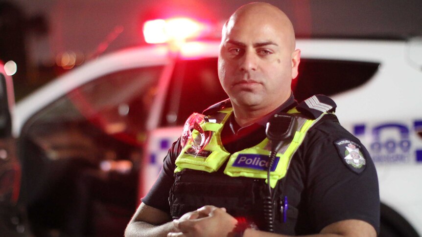 A police officer with a bald head, in his uniform, stands in front of a police car with its red light on.
