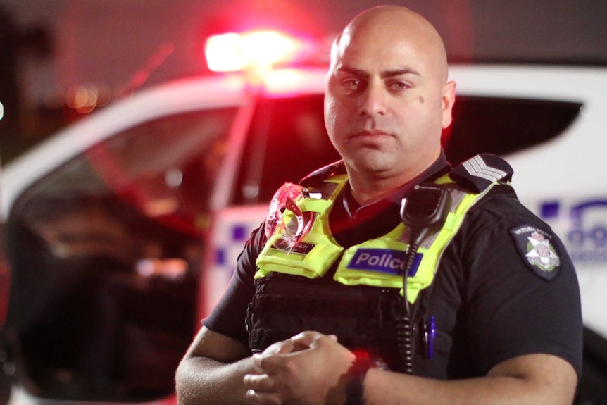 A police officer with a bald head, in his uniform, stands in front of a police car with its red light on.