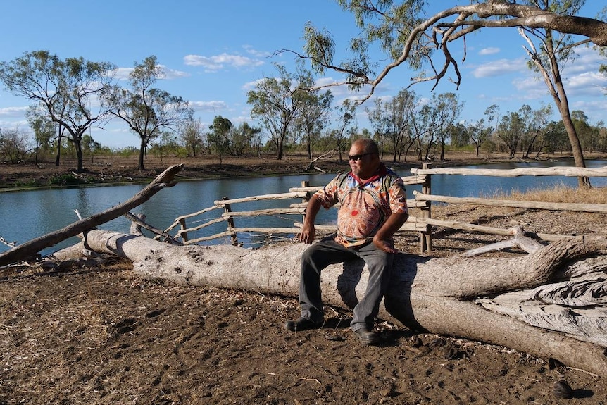 A man sits on a log next to a lagoon and stares into space