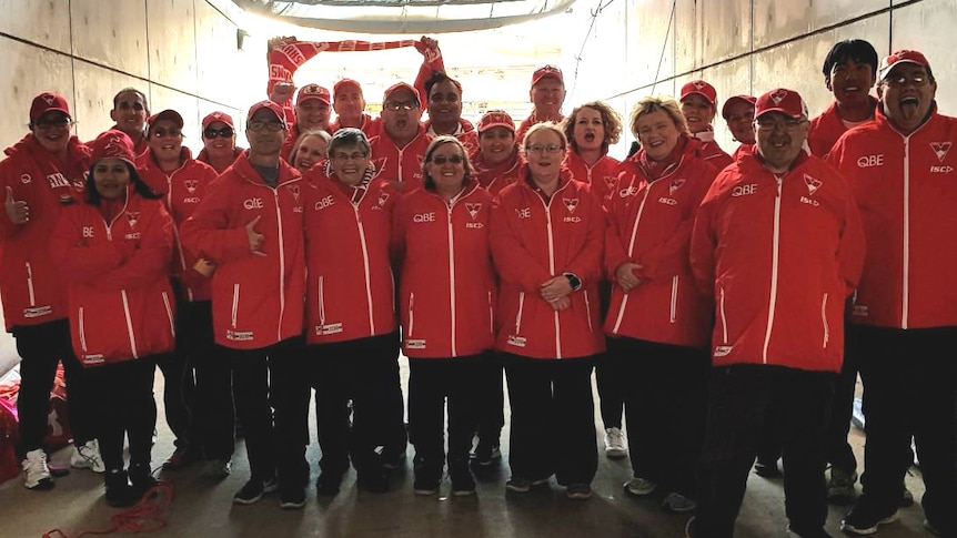 A group of about 20 people stand facing the camera wearing red and white jackets and hats