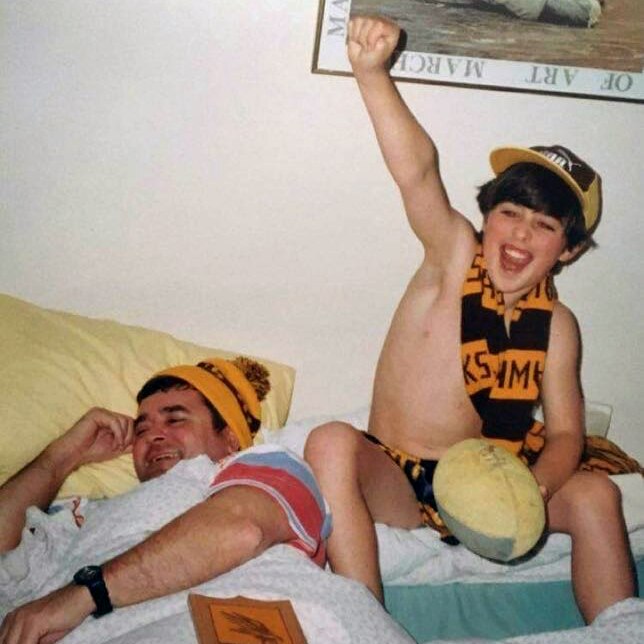 A boy sits wearing a Hawks AFL scarf cheers holding up his arm. A man lying in bed next to him smiles.