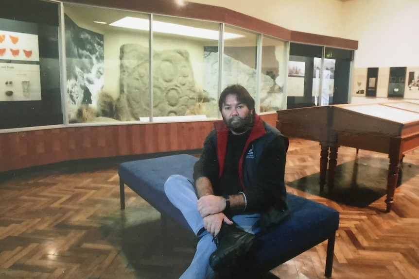 A man sits in on a bench in a museum, there is a stone on display behind him with circular carvings