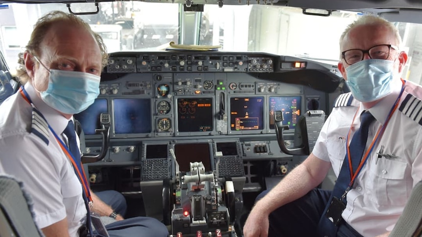 Two male pilots in the cockpit of a plane wearing masks.