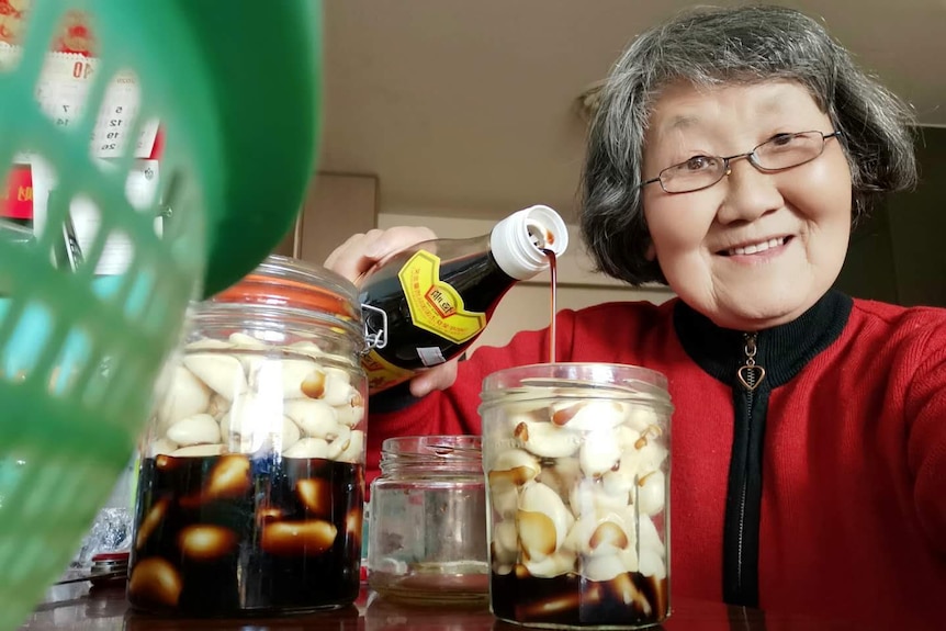 Weibin Xia smiles at the camera while pouring soy sauce in a jar of garlic cloves. She has short grey hair and glasses.