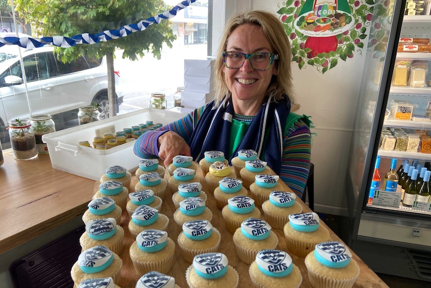 A smiling woman holds a large tray of cupcakes with the Cats AFL team logo on them.