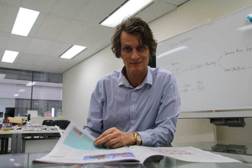 Mr Van Ek is wearing a patterned shirt, with a newspaper in front of him and a whiteboard in the background.