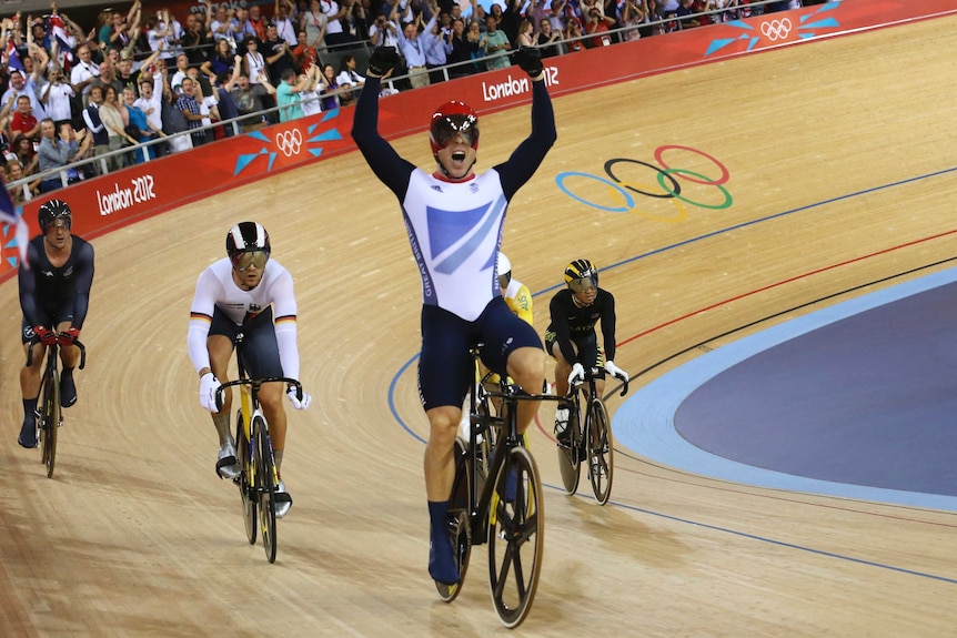 A victorious British Olympic track cyclist roars with his arms in the air at the finish line.