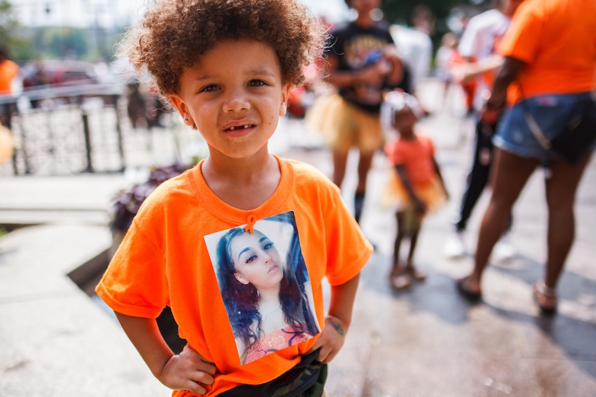 A young boy in an orange t-shirt holding a photo of his mother