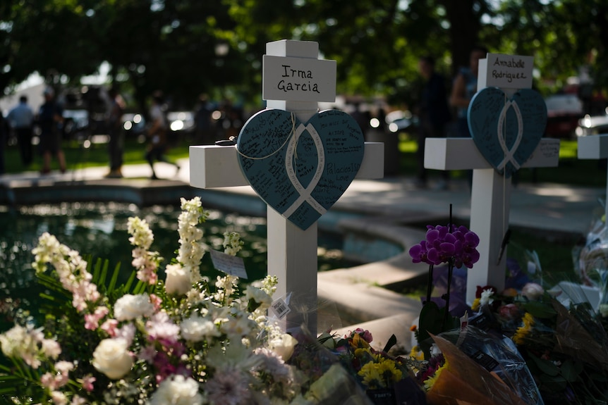 the name Irma Garcia is written on a white card above a blue heart placed on a white cross with flowers placed below