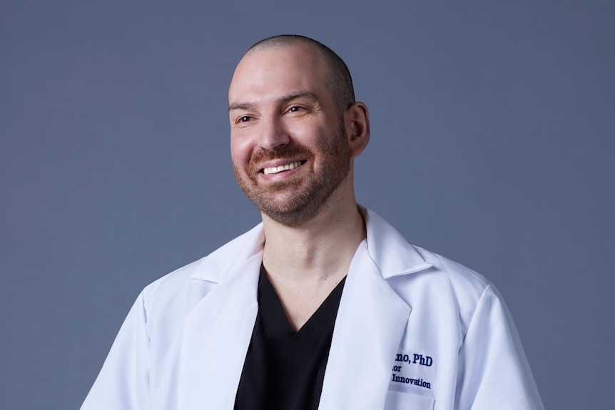 A bald man with stubble in a monogrammed lab coat over a navy tshirt smiles