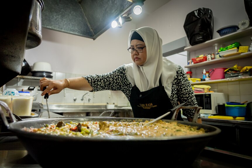 A woman wearing a hijab stands over a large wok and is mixing the food with a wooden spoon.