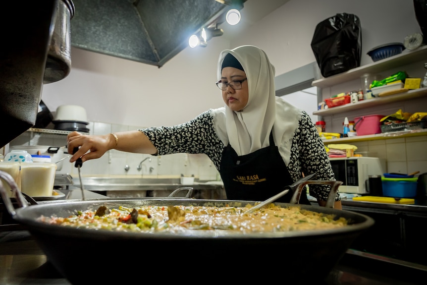 A woman wearing a hijab stands over a large wok and is mixing the food with a wooden spoon.