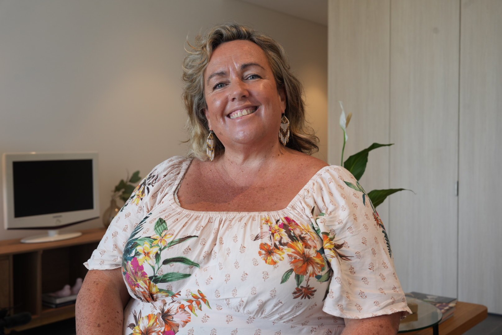 A woman with curly blonde hair wearing a floral top smiles inside her rental with a peace lilly behind her.