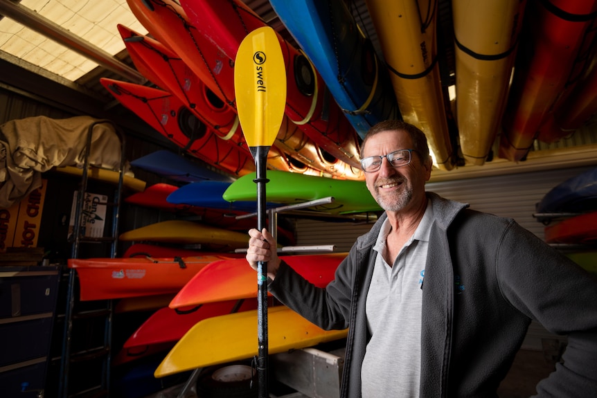 Kym Werner smiles as he holds a canoe paddle in front of stacked up canoes