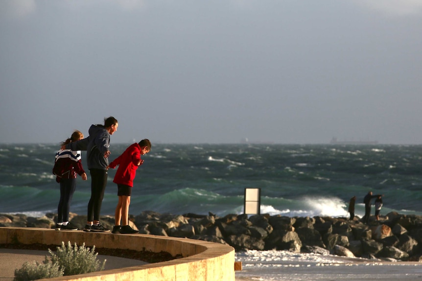 Three children lean into the wind with their arms outstretched as a storm approaches City Beach