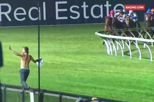 A shirt-less man holds up his arms as horses run around the track in the background