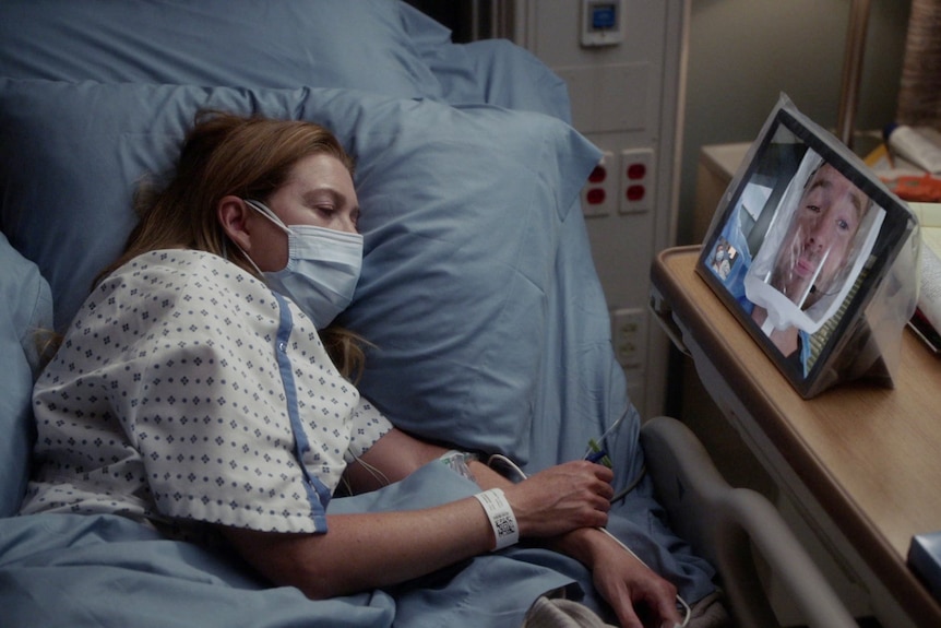 A woman lays on her side in a hospital bed as she face times a man. 