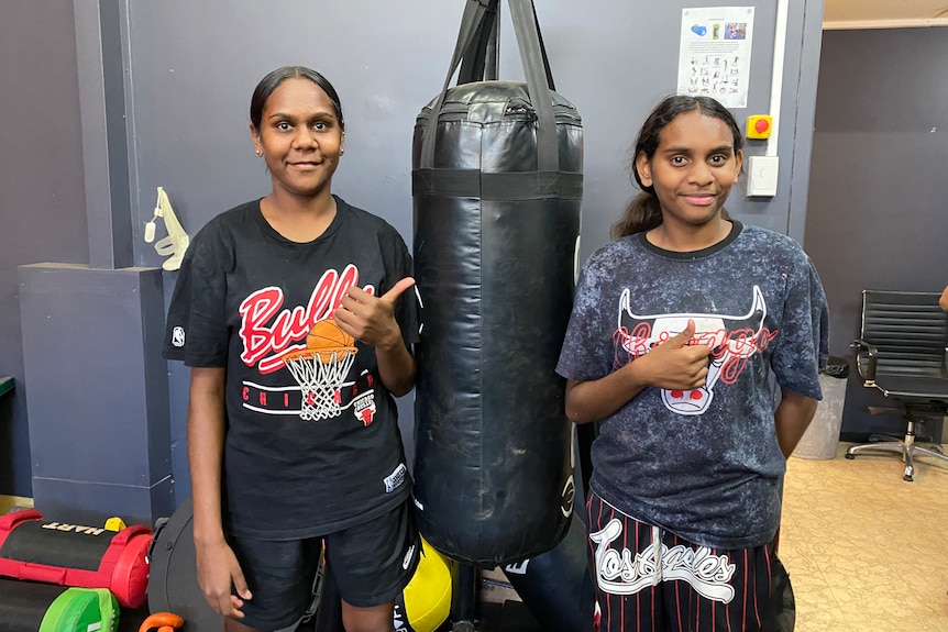 Two young girls stand on either side of a back boxing bag and smile at the camera with thumbs up.