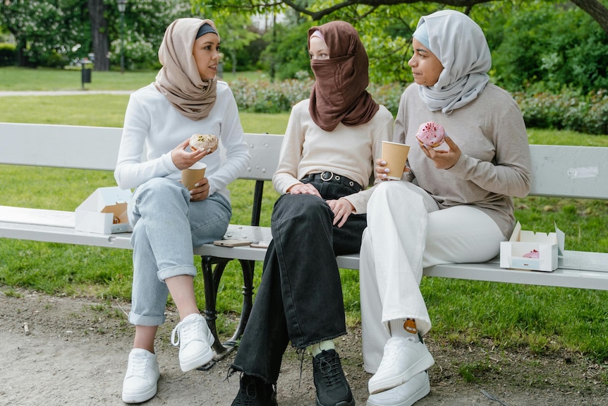 Three women sit on a wooden bench eating donuts wearing jeans and sneakers.. One covers her face with a niqab. All wear hijabs.