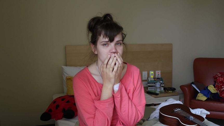 A young woman sitting on a hospital bed in pink pyjamas with her hands over her mouth and a resigned look on her face.