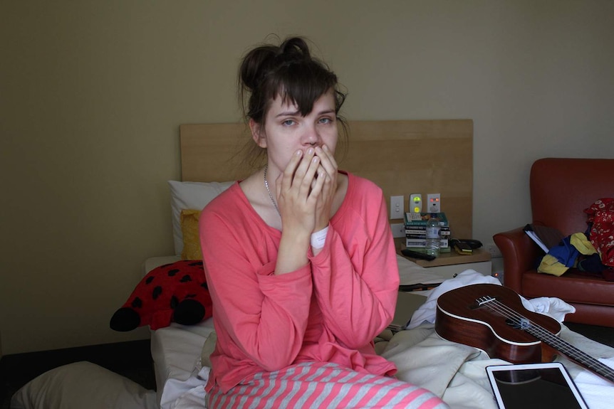 A young woman sitting on a hospital bed in pink pyjamas with her hands over her mouth and a resigned look on her face.