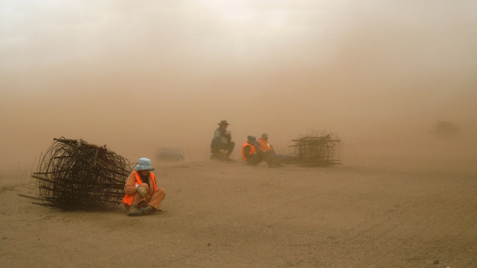 A group of people in high visibility hiding behind a roll of fencing wire as a storm of red dust passes over them.