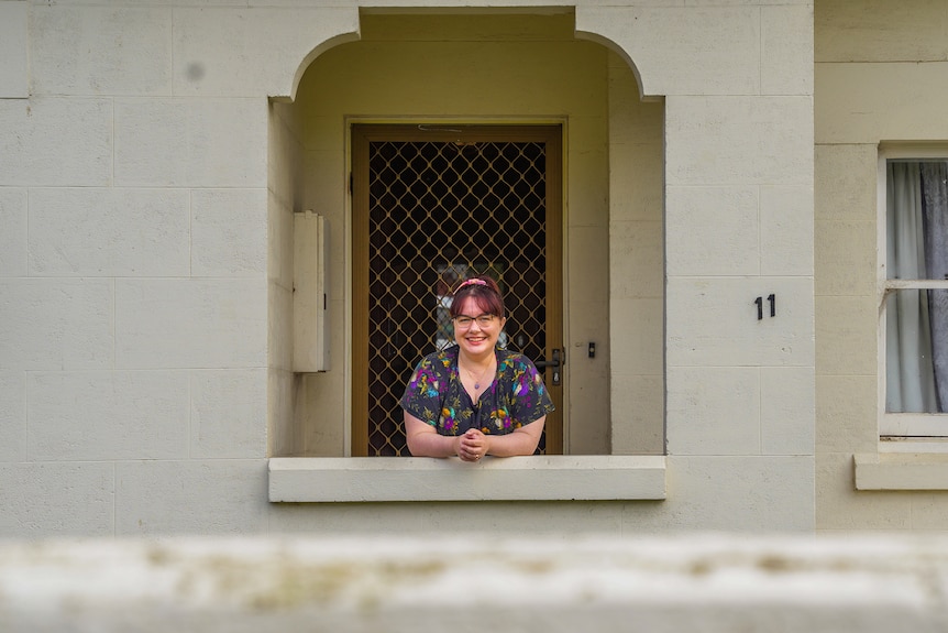 A woman with glasses and a floral dress leans against a door frame smiling.