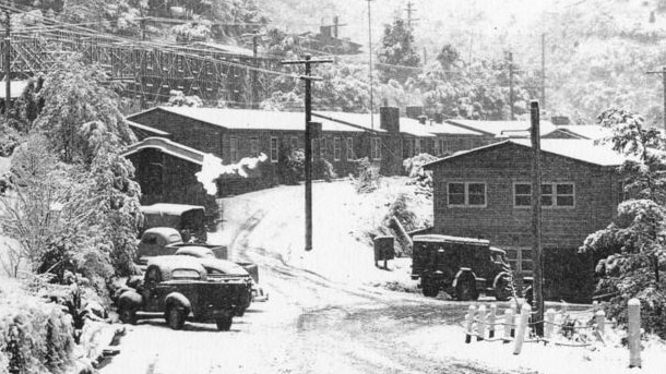A black and white image of a snow-capped street in a village with 1940s style cars parked outside cabins.