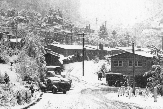 A black and white image of a snow-capped street in a village with 1940s style cars parked outside cabins.