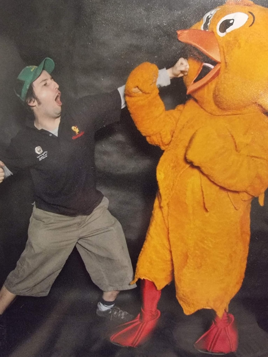 A man in shorts and a black tshirt with the Chickenfeed logo on it is pretending to punch a giant yellow chicken mascot