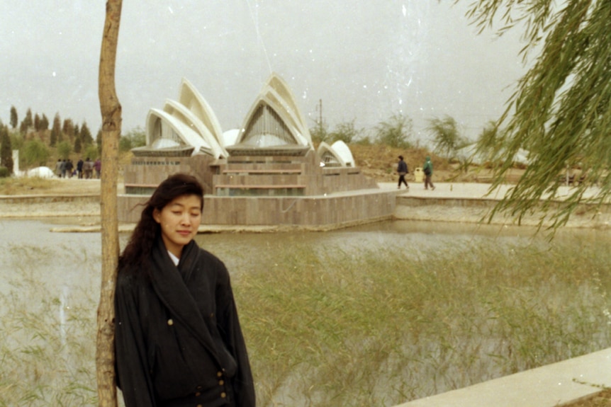 You view a woman with her eyes closed posing next to a tree with a fake Sydney Opera House behind her.