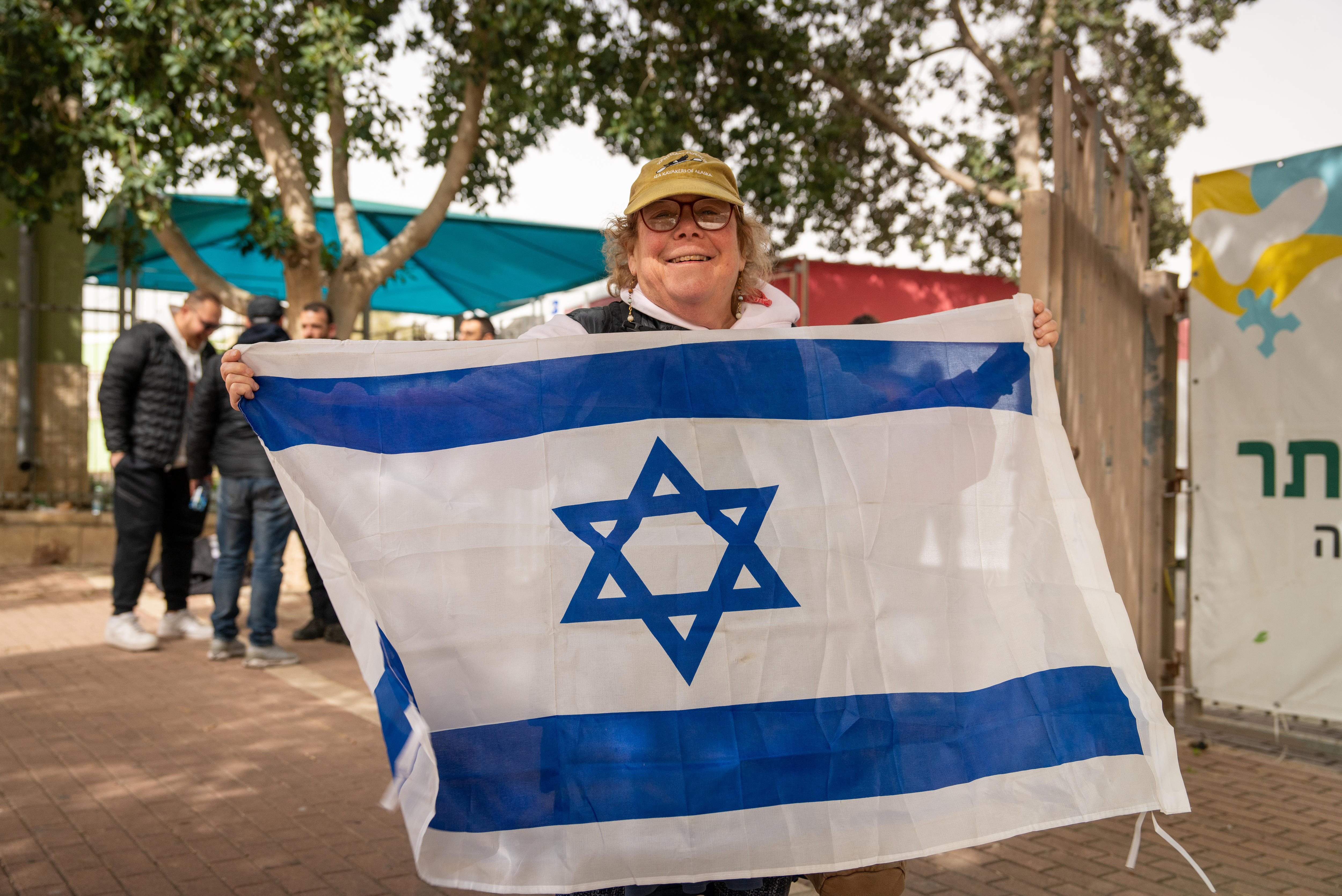 Foto de uma mulher de 50 anos, cabelo loiro curto, usando boné verde, óculos vermelhos e segurando uma bandeira israelense.