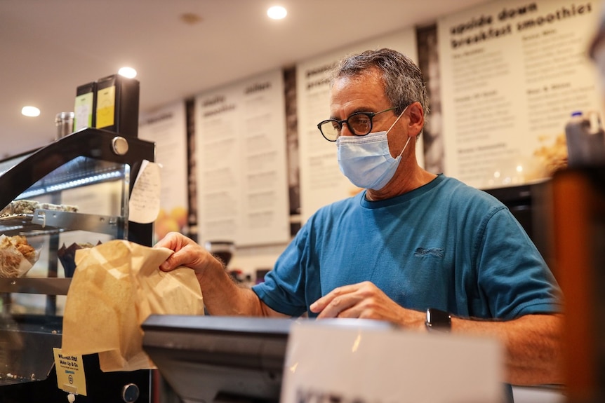 A man wearing a mask serves food in a brown paper bag in a cafe.