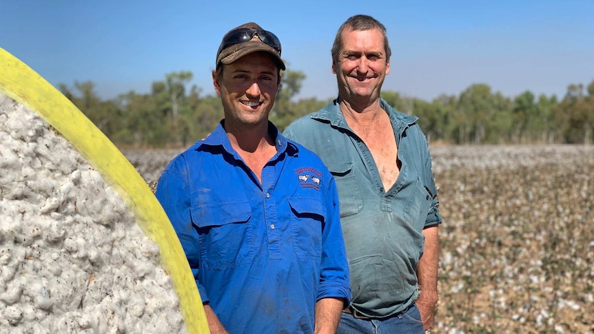 two men standing next to a cotton bale and a cotton crop.