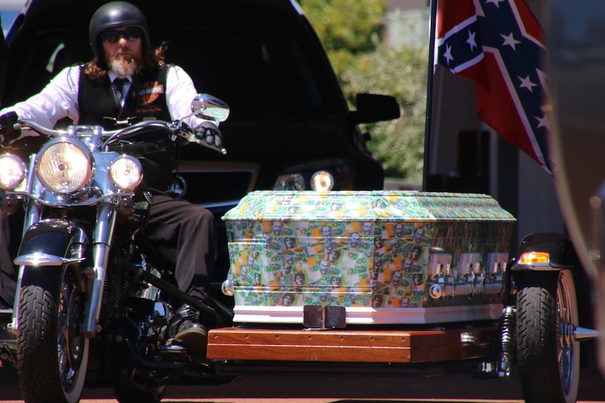 A coffin, printed with $100 bills, sits next to a man on a Harley.