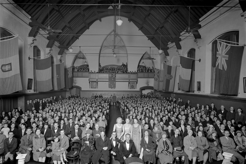 A large audience at Perth Town Hall looks towards the stage, flags surround the room