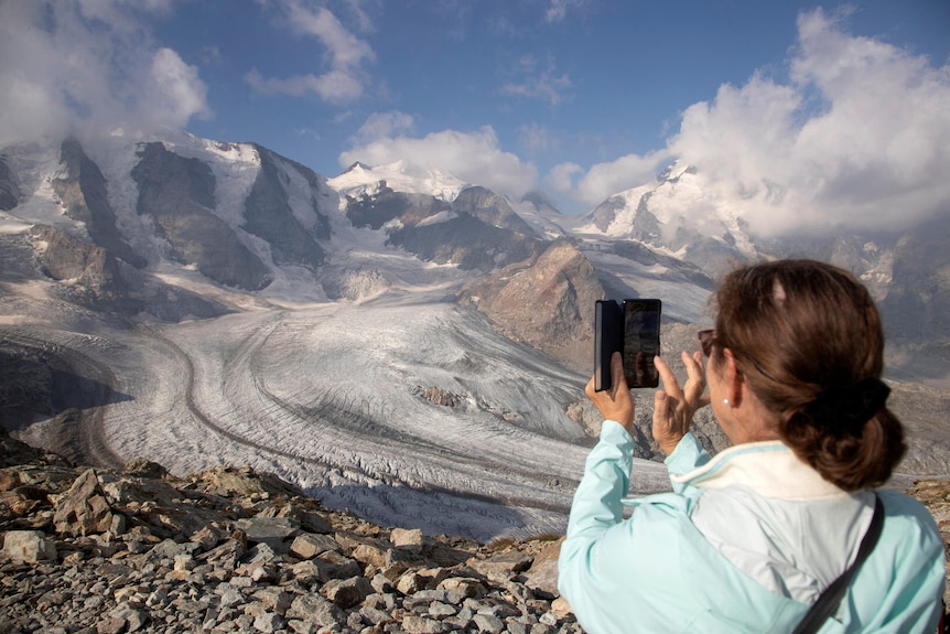 A woman in a blue jacket takes a photo of the Swiss alps with her mobile phone. 