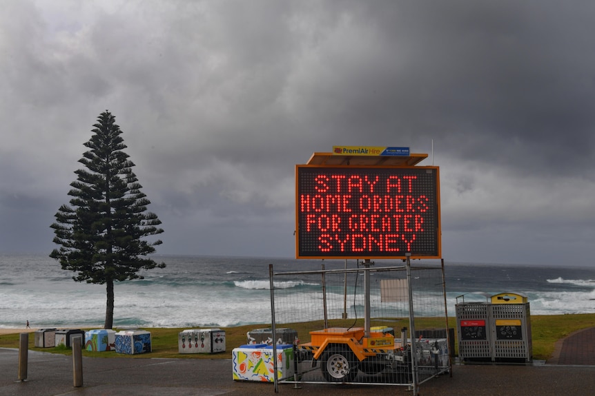 A digital road sign on Bondi Beach displays "Stay at home orders for Greater Sydney" on a gloomy overcast day