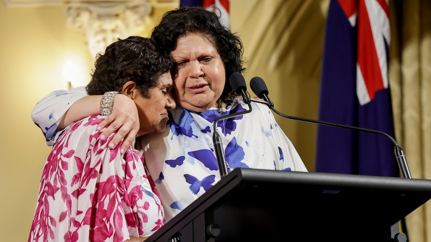 Mechelle Turvey hugs another woman on stage during the WA Australian of the Year awards ceremony, with flags off to their left.