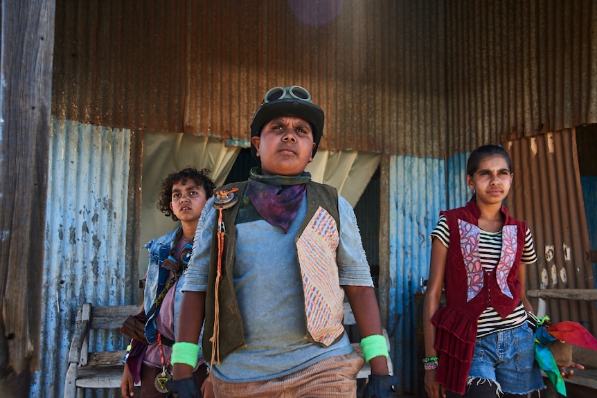 Three kids stand in front of rusting and weathered corrugated iron building and look into distance with serious expressions.