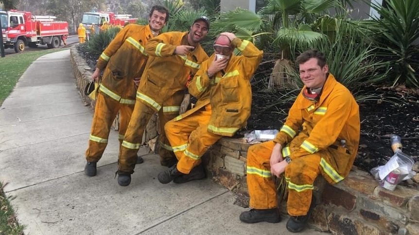 four men in yellow suits laugh at the camera with red trucks in the background.