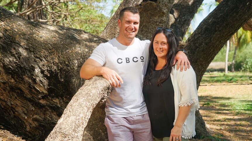 A man and woman stand in front of a tree at a park with their arms around each other. They're smiling.