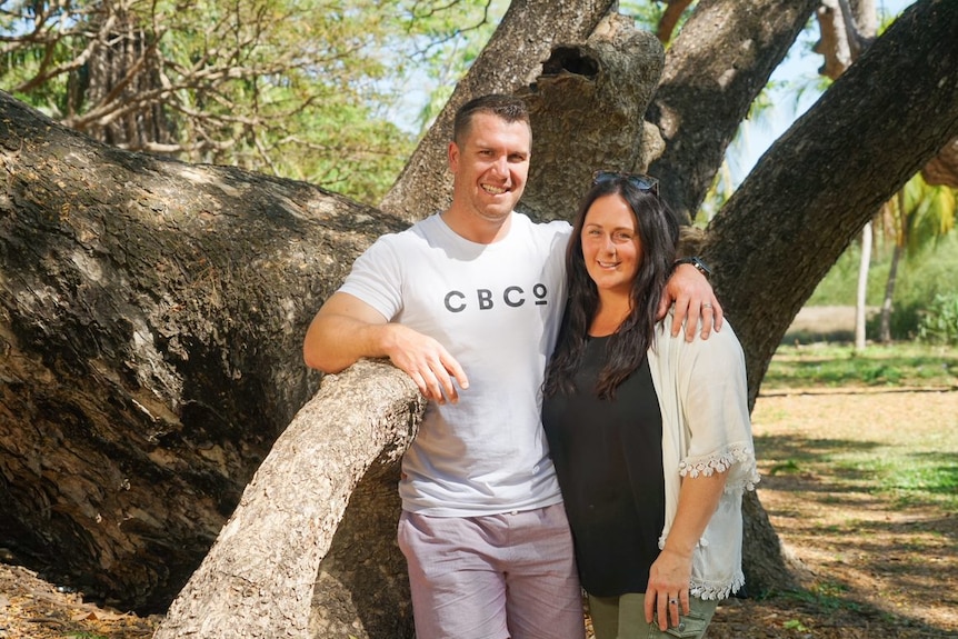 A man and woman stand in front of a tree at a park with their arms around each other. They're smiling.