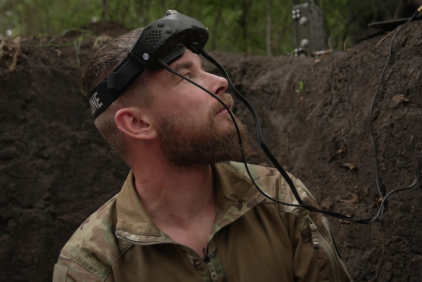 A close up of a man with a full beard, wearing camouflage gear and a headset looking out of a fox hole.