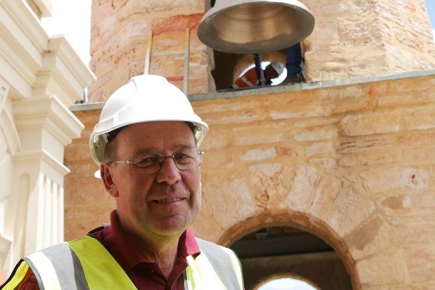 A man in a hard hat and hi-vis stands in front of a church. A bell is being hoisted skyward on a crane.