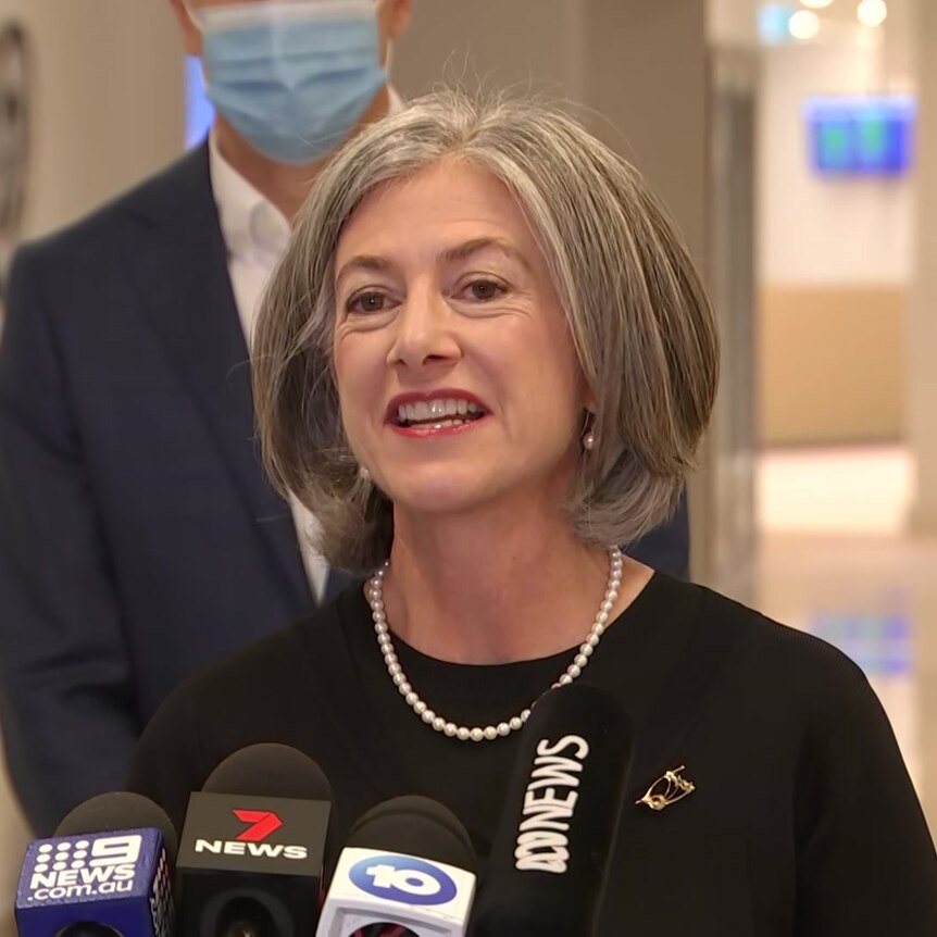 A smiling woman stands in a hospital corridor with media microphones in front of her