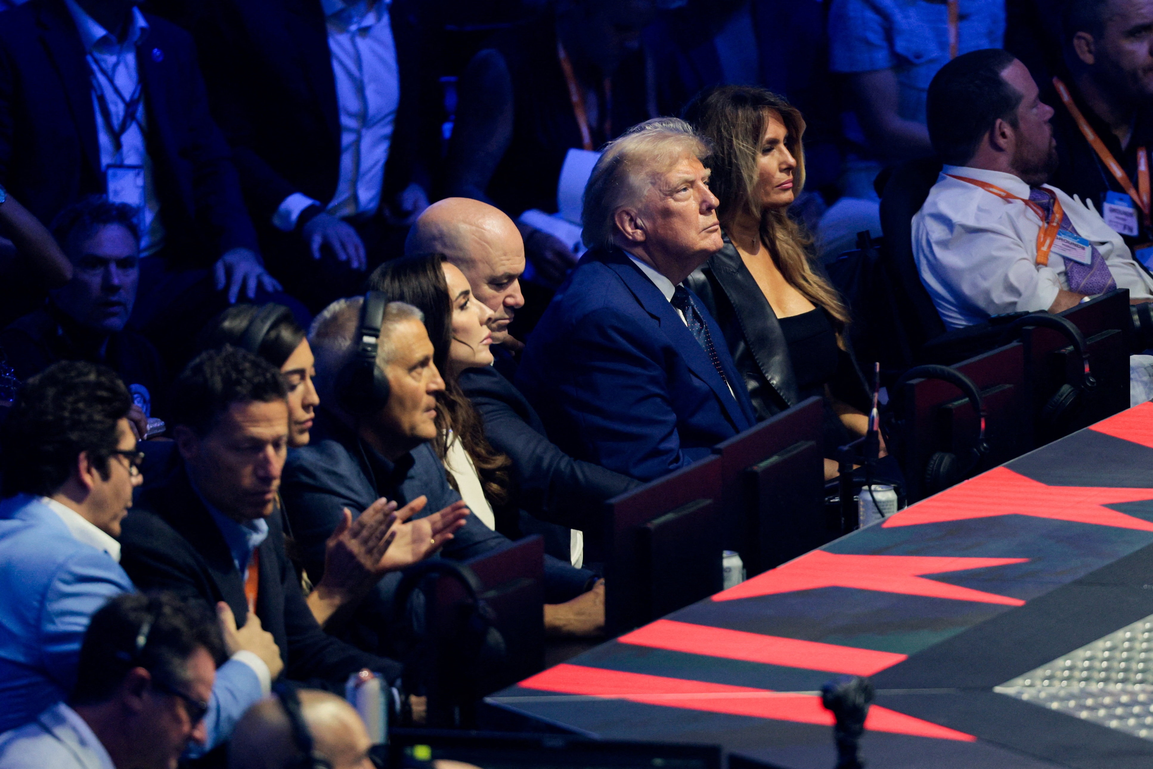 Trump and Melania sit ring side among other people, red lights before them.