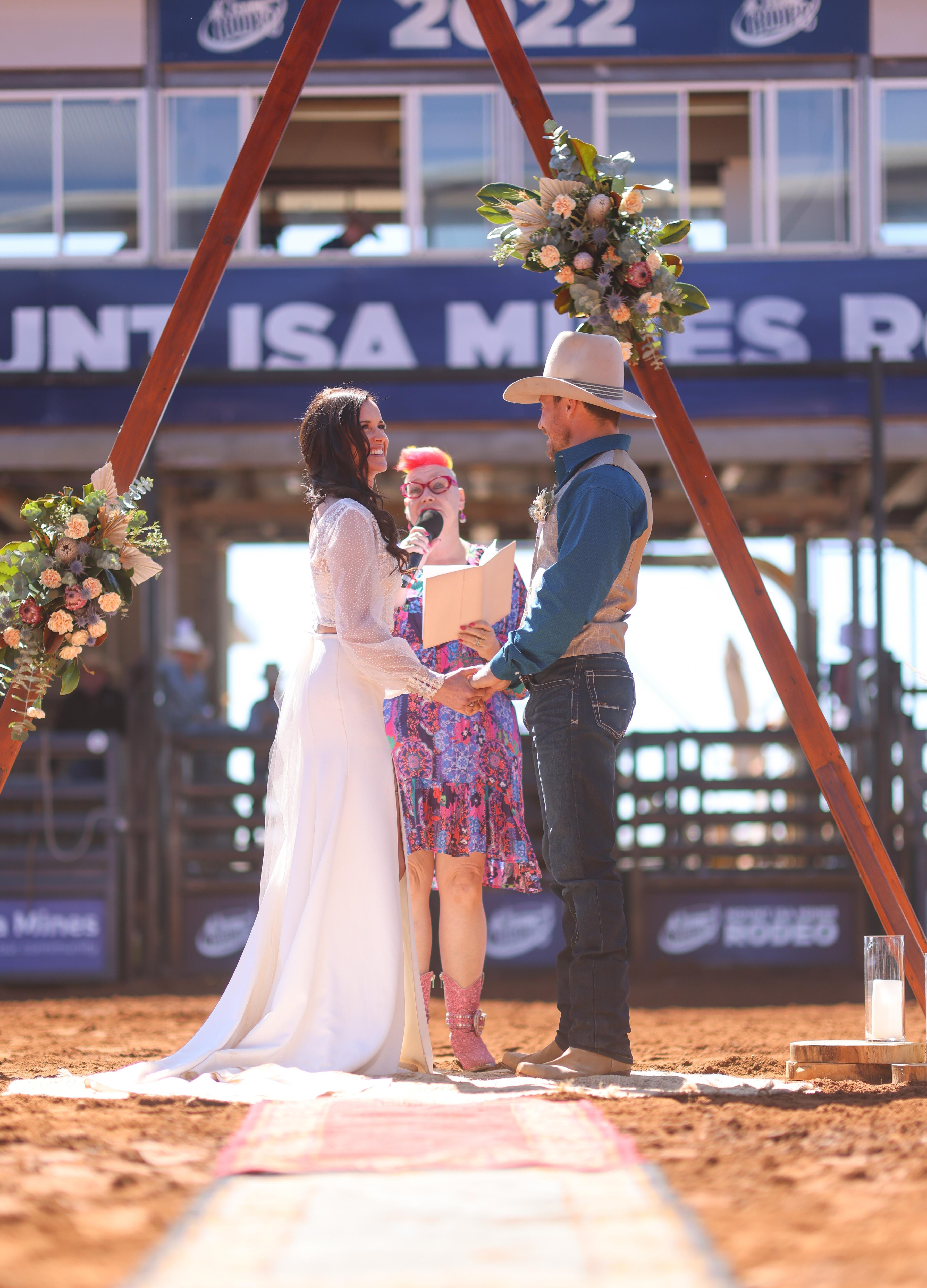A bride and groom stand at a triangle timber alter on red dirt ground with a colorfully dressed celebrant holding a microphone.