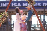 A bride and groom stand at a triangle timber alter on red dirt ground with a colourfully dressed celebrant holding a microphone.