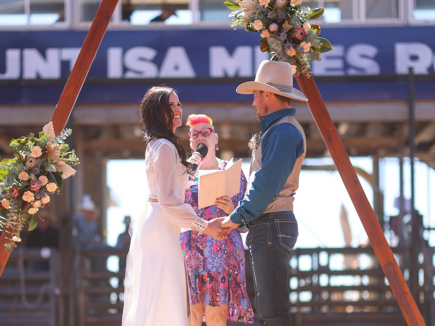 A bride and groom stand at a triangle timber alter on red dirt ground with a colourfully dressed celebrant holding a microphone.